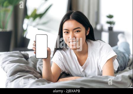 Excitée jolie fille asiatique se trouve dans la chambre sur le lit, montre un smartphone avec un blanc écran maquette, vide espace pour votre publicité ou présentation, regardant l'appareil photo, souriant heureux Banque D'Images