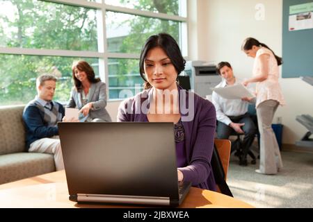 Photo d'une femme assise sur une table dans une salle du personnel très occupée travaillant sur un ordinateur portable au bureau. Banque D'Images