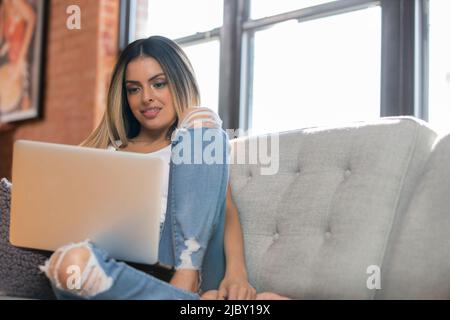 Prise de vue sous angle d'une femme hispanique se prélassant sur un canapé gris avec ordinateur portable Banque D'Images