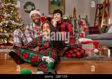 Joyeux portrait de vacances d'un couple de race mixte avec leur jeune fille regardant dans l'appareil photo souriant avec l'arbre illuminé en arrière-plan. Banque D'Images