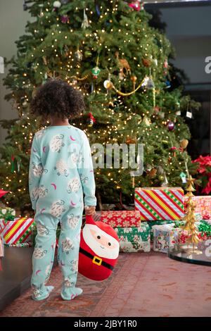 Petite fille en onesie pyjama debout et regardant l'arbre de Noël dans les merveilles. Banque D'Images