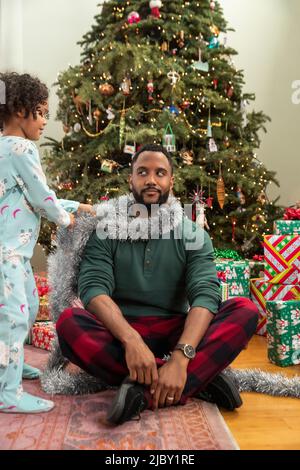 Une petite fille décorant son père avec guirlande devant l'arbre de Noël. Banque D'Images