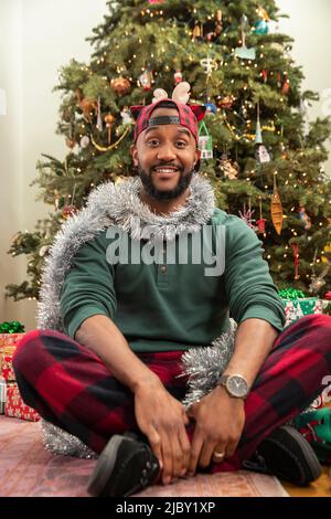 Portrait d'un Africain américain assis devant un arbre de Noël dans son pyjama décoré de guirlande argentée Banque D'Images