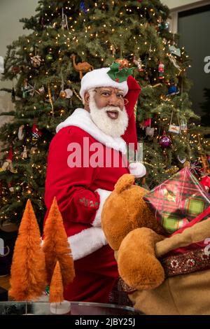 Un Père Noël surpris regardant derrière son épaule à la caméra tout en plaçant des cadeaux sous l'arbre de Noël. Série Père Noël noir. Banque D'Images