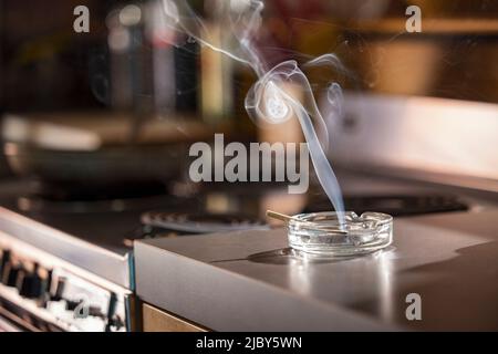 Maison intérieur, détail du cendrier qui a un joint de pré-rouleau de CBD éclairé, cigarette avec la fumée s'élevant assis dans la cuisine à côté du dessus de cuisinière Banque D'Images