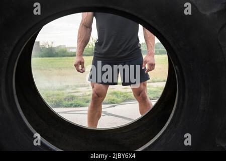 Vue d'un homme hispanique musclé portant un débardeur à travers le pneu du tracteur qu'il a retourné. Banque D'Images