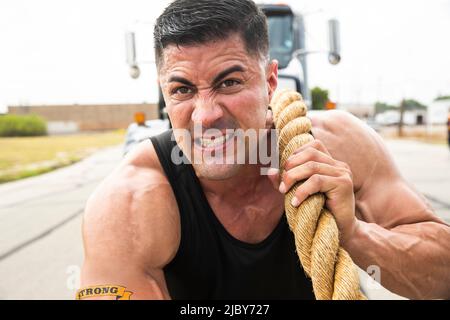 Un hispanique musclé portant un débardeur avec un tatouage « fort » sur son biceps, qui se met à tirer un semi-camion avec un grand câble de remorquage Banque D'Images