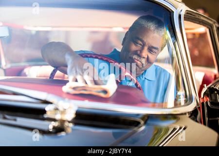 Homme plus âgé assis dans le siège conducteur de la berline sport Super 88 d'Oldsmobile 1960, essuyant le tableau de bord avec un chiffon anti-poussière Banque D'Images