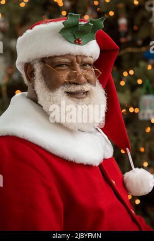 Un homme afro-américain habillé comme le Père Noël assis devant un arbre de Noël regardant un appareil-photo souriant. Banque D'Images