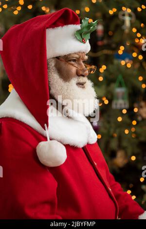 Un homme afro-américain habillé comme le Père Noël assis devant un arbre de Noël a tourné le profil, regardant hors de la caméra. Banque D'Images