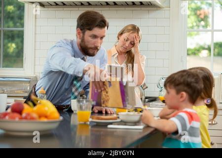 Maman avec sa famille dans la cuisine, luttant pour prendre le petit déjeuner pour les enfants et prêt à commencer la journée. Vérification de la connexion de la tablette avec le médecin Banque D'Images