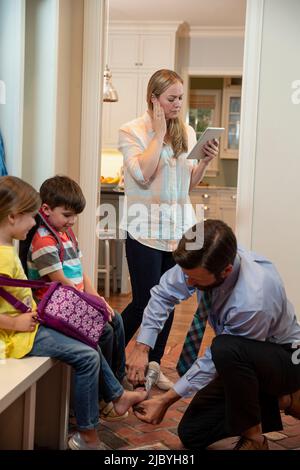 ly le matin se mettre, papa monter des chaussures pour enfants dans les mudroom avant de partir à l'école, maman parlant avec le médecin sur iPad décrivant ses symptômes Banque D'Images