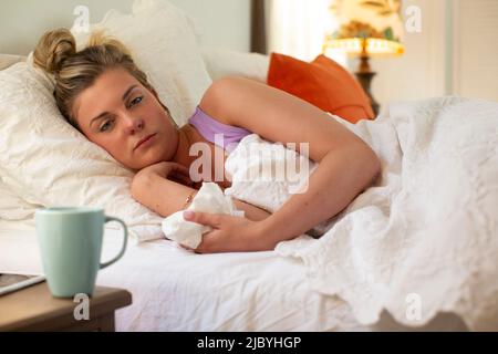 Jeune femme dans son lit discutant de sortir du lit, tenant le papier de soie dans la main, smartphone et tasse de café assis sur une table de chevet Banque D'Images