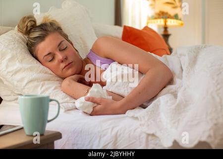 Une jeune femme dormant dans son lit, tenant le papier de soie à la main, un smartphone et une tasse à café assis sur une table de chevet Banque D'Images