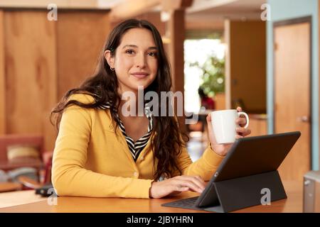 De jeune femme ethnique portant un chandail jaune avec un chemisier rayé noir et blanc, assis au bar dans la cuisine du centre-ville loft avec iPad et tasse de café Banque D'Images