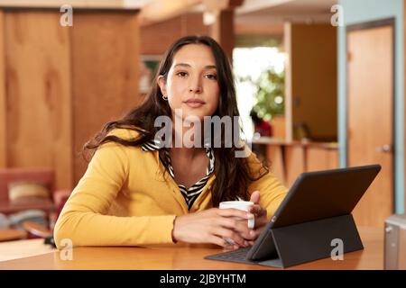 De jeune femme ethnique portant un chandail jaune avec un chemisier rayé noir et blanc, assis au bar dans la cuisine du centre-ville loft avec iPad et tasse de café Banque D'Images