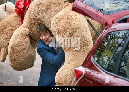 3/4 MCU d'un jeune homme saisissant un grand ours en peluche du coffre de sa voiture, le jetant par-dessus son épaule et marchant hors de la caméra. Banque D'Images