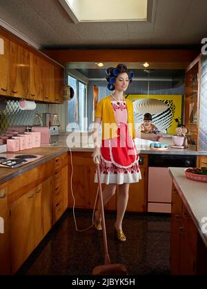 Femme passant l'aspirateur dans la cuisine tout en portant des rouleaux à cheveux dans une cuisine de style du milieu du siècle. Banque D'Images