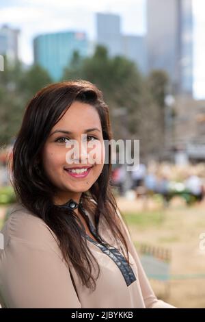 Portrait souriant d'une jeune femme ethnique avec de longs cheveux noirs assis dans le parc, en regardant par-dessus l'épaule vers l'appareil photo Banque D'Images