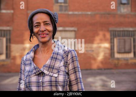 Portrait d'une femme plus âgée portant un chapeau en tricot debout dans une allée souriante regardant l'appareil photo, mur de briques en arrière-plan Banque D'Images