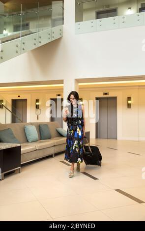 Hispanic woman with suitcase using cell phone marcher dans le hall de l'hôtel Banque D'Images