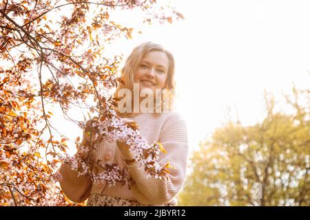 Portrait d'une jeune femme aux cheveux équitables portant un gilet rose, robe debout près d'un oiseau-cerisier en fleurs dans le parc. Banque D'Images