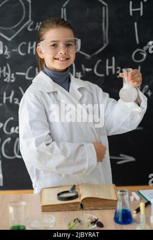 écolière en manteau blanc faisant des expériences avec des liquides en laboratoire de chimie, fond de tableau noir de l'école avec des formules scientifiques dessinées à la main, à Banque D'Images