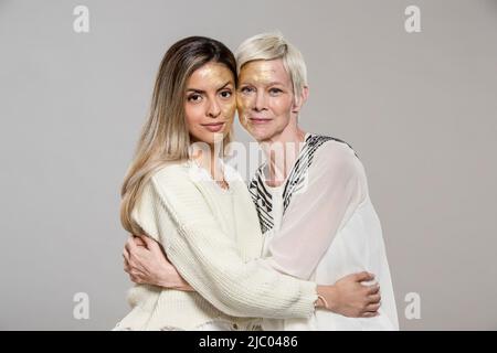 Portrait de femmes caucasiennes et hispaniques avec des visages peints en or embrassant sur fond gris. Banque D'Images