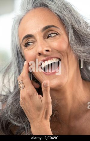 Femme d'âge moyen avec cheveux gris brosses cheveux hors de son visage et regarde hors de l'appareil-photo rire. Banque D'Images