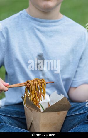 Enfant méconnaissable mangeant des bâtonnets de nouilles. La sauce soja sale se tache sur un t-shirt blanc. Plats à emporter. À l'extérieur Banque D'Images