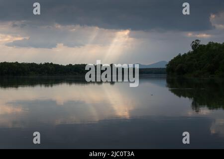 Rayons de soleil perçant à travers les nuages au-dessus de la rivière, forêt sur la rive de la rivière et silhouette de montagne à distance Banque D'Images