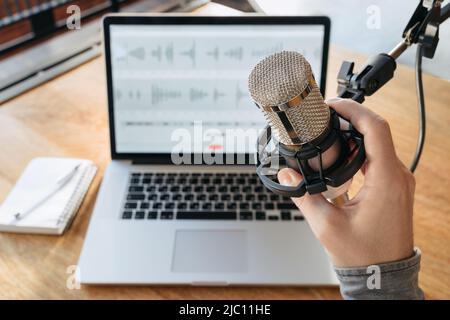 Gros plan de la main de l'homme tenant le microphone dans le studio de radiodiffusion. Intérieur du podcast du studio d'accueil. Ordinateur portable, stylo et bloc-notes sur une table en arrière-plan. Création d'un podcast dans le studio de podcast domestique. Vue première personne Banque D'Images