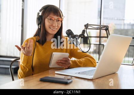 Magnifique baladoteur femelle dans le casque et les lunettes gestant tout en diffusant le podcast audio en studio, en interviewant un gest, regardant vers. Une jeune femme blogueuse parle dans un micro Banque D'Images