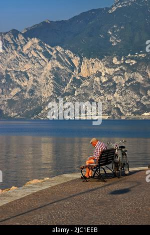 Un cycliste à capuchon plat, un chemisier à carreaux et un short lit le journal sur le banc de bord de mer de Lago di Garda à Torbole sul Garda, Trentin-Haut-Adige, en Italie, alors que la lumière du soleil tôt le matin éclaire les pentes calcaires des pics pré-alpins de Cima sur le côté lointain du lac. Banque D'Images