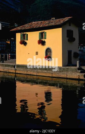 Les murs ocre jaunes de la casa photogénique ou Casetta del Dazio, un ancien poste de péage ou de douane autrichien du début du 18th siècle, brillent sur un fond plus sombre à l'entrée du port de Torbole sul Garda, Trentin-Haut-Adige, Italie, Comme ils se reflètent dans le soleil d'été tôt le matin dans les eaux doucement ondulant du lac de Garde. Banque D'Images