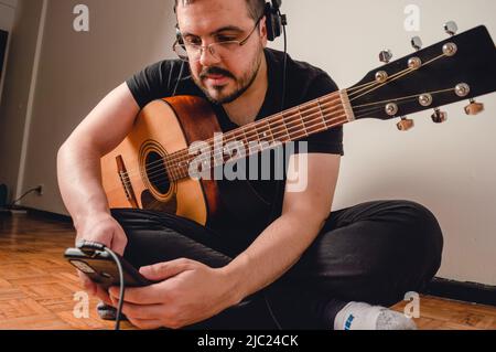 jeune latino-caucasien vénézuélien, assis sur le plancher du salon de sa maison, avec sa guitare acoustique pratiquant en regardant la vidéo tutoria Banque D'Images