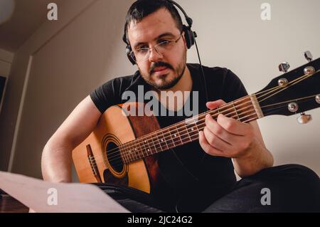 jeune homme du caucase du sud avec une barbe, des écouteurs et des lunettes, vêtu de noir et assis sur le sol du salon à la maison, pratiquant la guita Banque D'Images