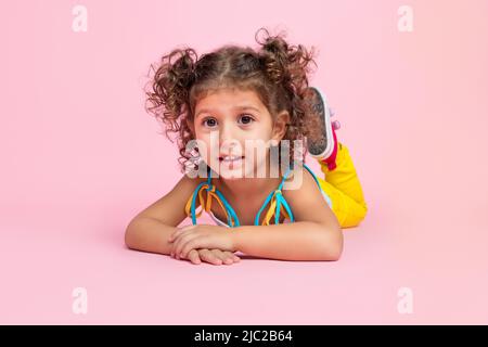 Portrait d'une jolie jeune fille aux cheveux ondulés et gaies, couché sur le sol et reposant l'été, isolée sur un fond rose pastel Banque D'Images