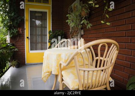 Ancienne maison de 1927 briques rouges avec porte d'entrée jaune et véranda en bois gris avec fauteuils en rotin et table en été. Banque D'Images
