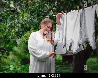 femme caucasienne d'âge moyen en robe blanche faisant des devoirs, pendant des vêtements sur la corde à linge dans la rue dans la cour de la maison de village, concept de jour de blanchisserie Banque D'Images