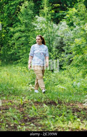 Portrait d'une jeune femme gras avec de longs cheveux rouges bouclés marchant sur le chemin dans la forêt du parc parmi les arbres verts. Été, week-end. Banque D'Images