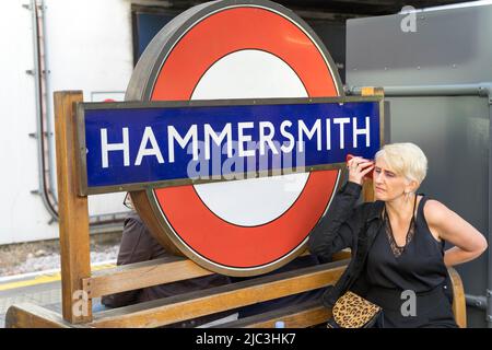 Femme passager en robe noire sur le téléphone se trouve à côté du panneau Hammersmith cocarde Londres Angleterre Royaume-Uni Banque D'Images