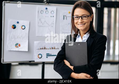 Portrait jeune adulte professionnel belle confiante femme caucasienne, chef de la direction, portant un costume d'affaires et des lunettes, personnalité amicale, debout dans un bureau moderne, regardant l'appareil photo, souriant Banque D'Images