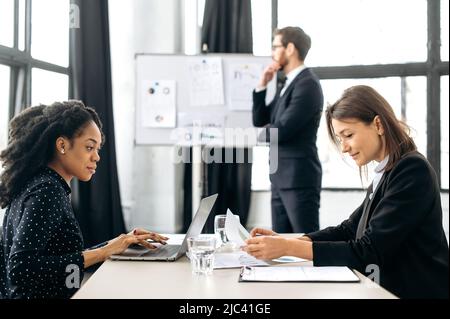 Des collègues féminins, une afro-américaine et une femme caucasienne, concentrés et prospères, sont assis à une table dans un bureau moderne, travaillent sur un projet, développent des start-up, communiquent avec les clients Banque D'Images