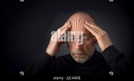 fatigué - 60 ans homme avec une barbe et des lunettes massant son front - une tête contre un fond noir avec un espace de copie Banque D'Images