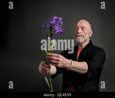 Tête et épaules portrait d'homme âgé à tête chauve et barbu dans un kimono en admirant ou en offrant une fleur d'iris, se concentre sur la fleur Banque D'Images