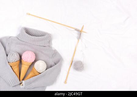 Chandail tricoté gris, aiguilles à tricoter en bois et cônes de crème glacée avec fil sur fond blanc. Tricotage, passe-temps et concept créatif fait main. Haut vi Banque D'Images