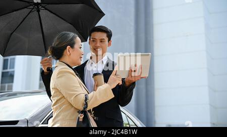 Femme d'affaires asiatique ou directrice générale qui a réussi à vérifier son travail sur une tablette numérique tout en se tenant à l'extérieur avec son assistant masculin sous la couchée Banque D'Images