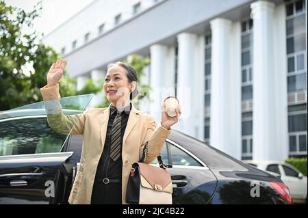 Une femme d'affaires asiatique agée et joyeuse qui sort de sa voiture de luxe salue ses collègues. Concept de style de vie urbain Banque D'Images