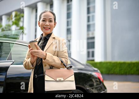 Portrait d'une belle femme d'affaires asiatique âgée ou d'une avocate professionnelle senior en costume formel sortant de la voiture, debout devant la comp Banque D'Images